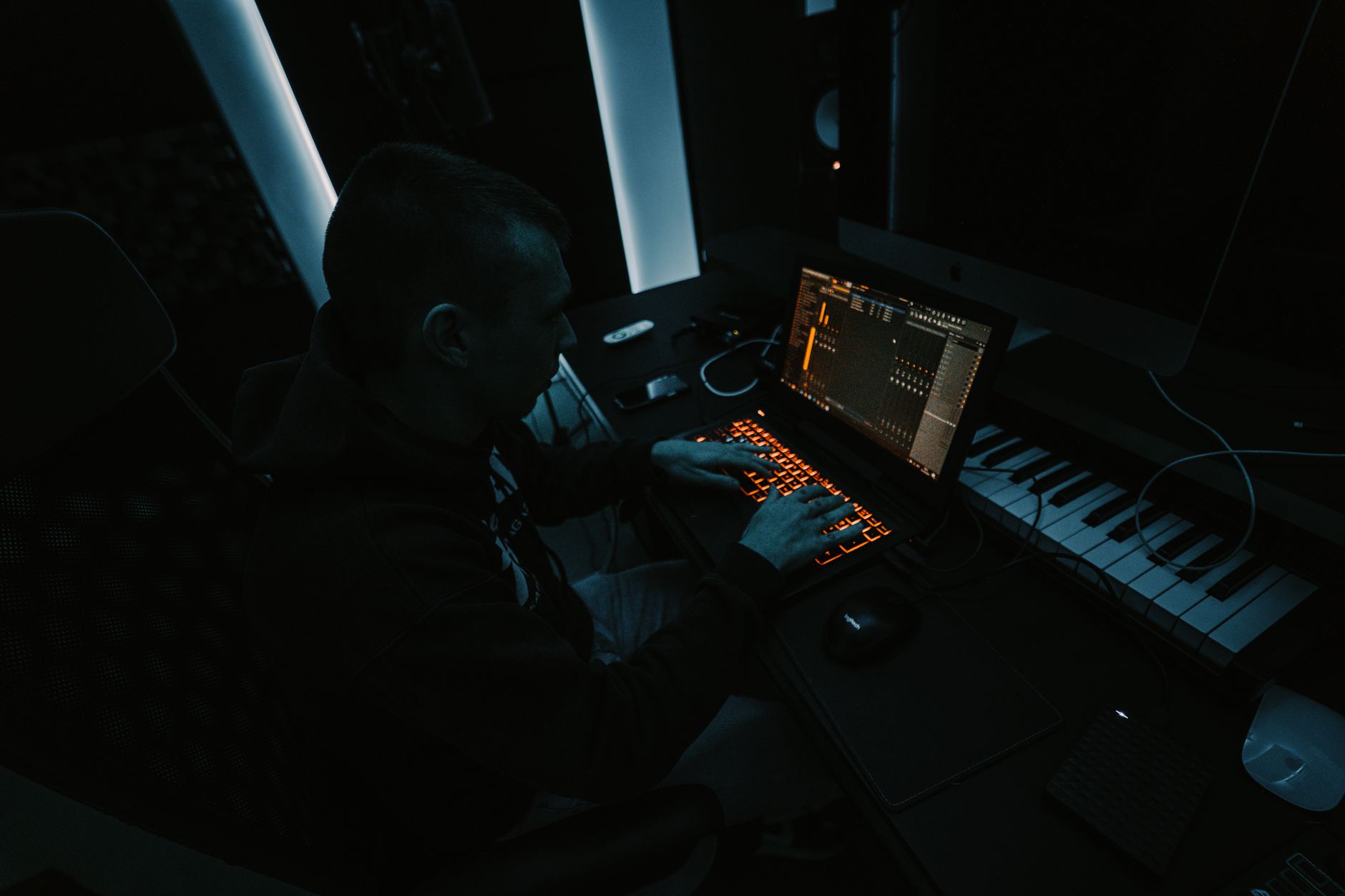 Music producer focused on composing tracks in a dimly lit studio with laptop and keyboard.