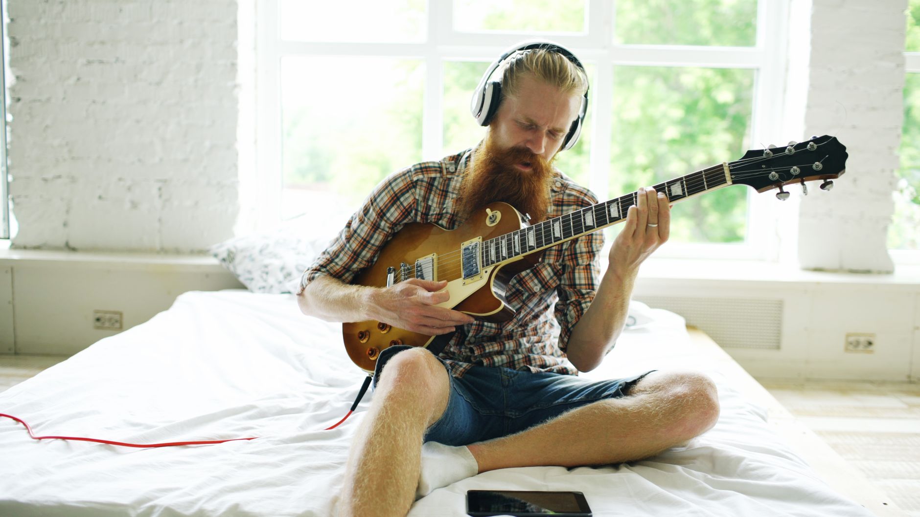 Bearded man in casual attire playing electric guitar with headphones indoors.