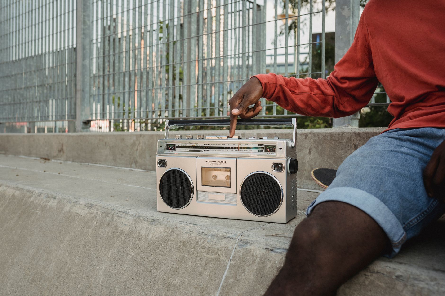 Crop anonymous black guy in denim shorts sitting on concrete step near metal railing and pressing button on old fashioned cassette recorder