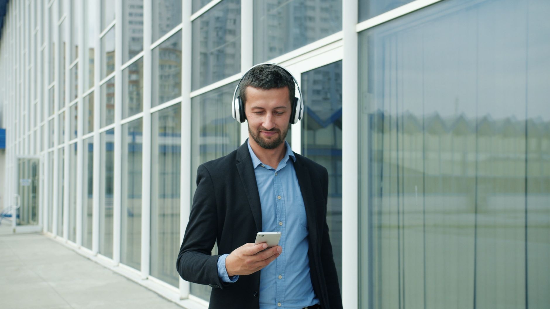 Smiling businessman listens to music with headphones outside a modern office building.