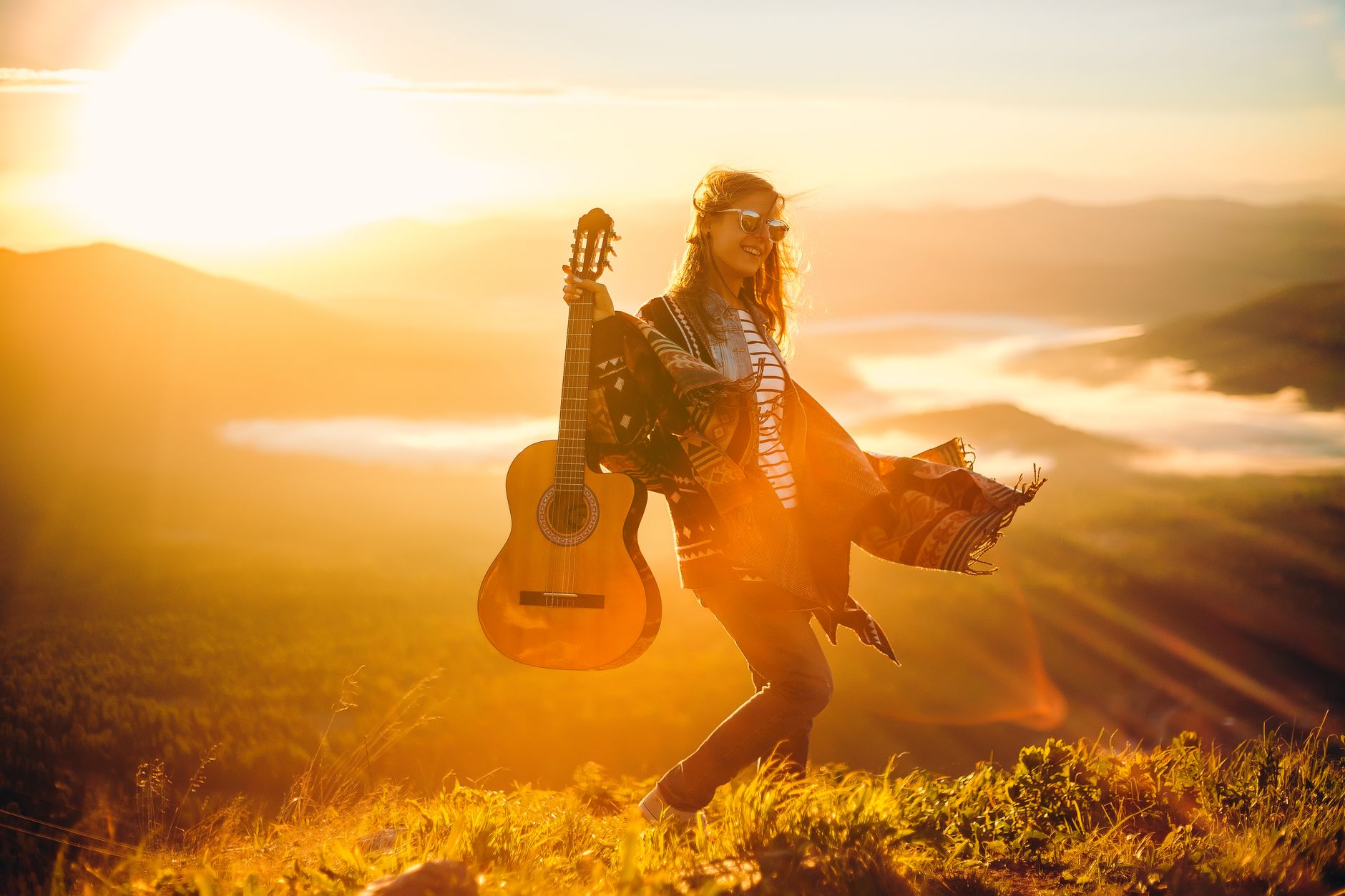 A woman stands in a grassy field with an acoustic guitar during a beautiful sunset, embracing the golden hour's warmth.