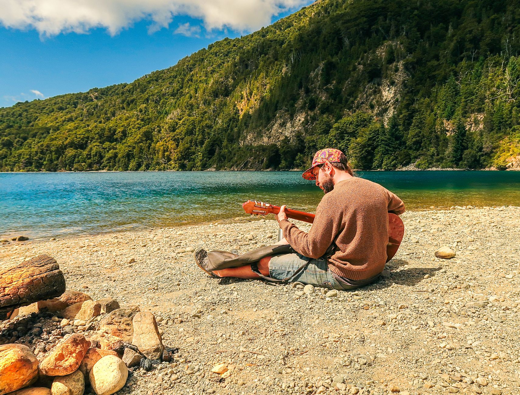 A man sits on a rocky beach playing guitar by a lake in Bariloche, Argentina.