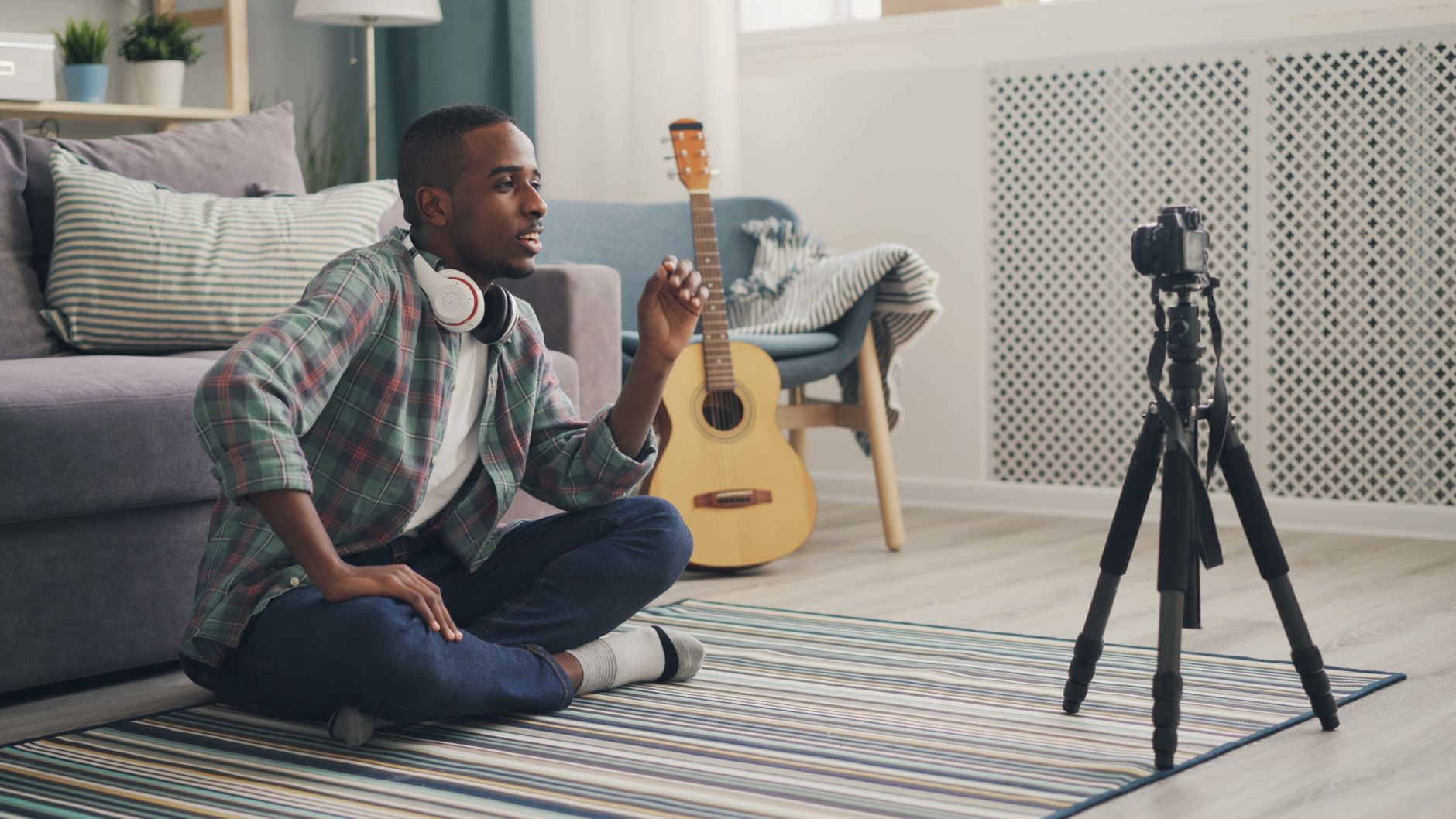 A young man wearing headphones records a video in a cozy home setting with guitar props.